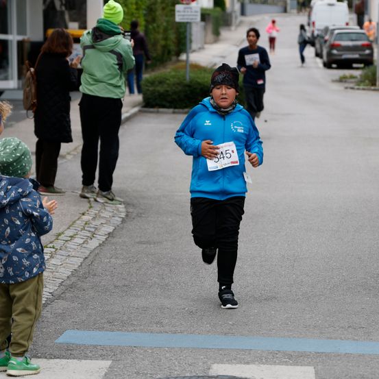 Ein junger Junge in einem blauen Jacke und schwarzen Hosen läuft in einem Rennen. Zuschauer beobachten vom Bürgersteig aus, und ein Kind steht auf der Straße. Autos sind entlang der Straße geparkt.