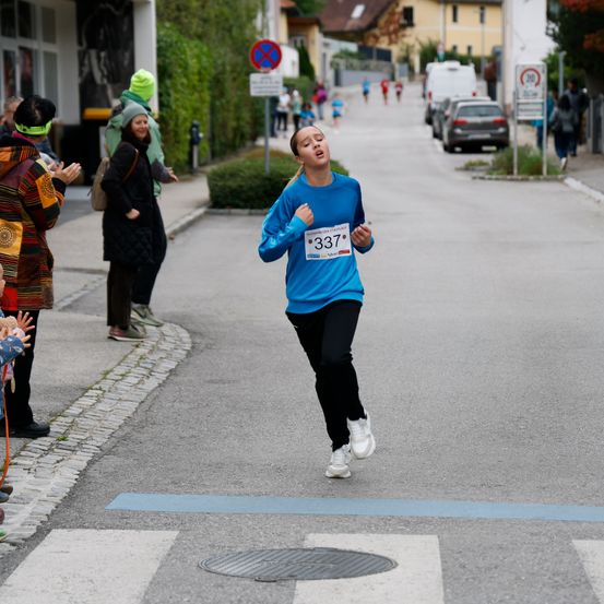 Eine Frau in einem blauen Shirt mit der Nummer 337 läuft bei einem Marathon. Zuschauer beobachten vom Bürgersteig aus.