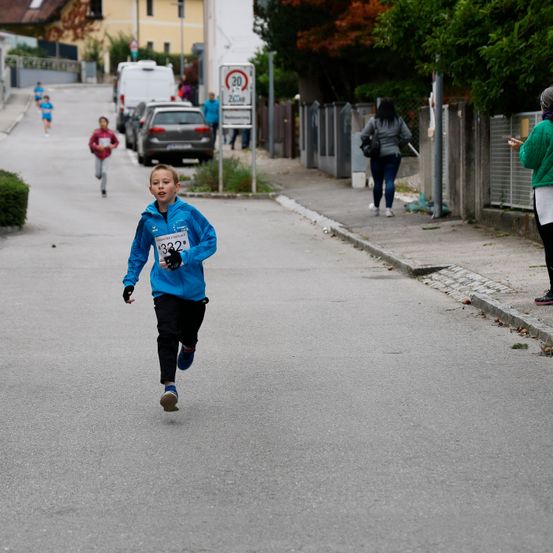 Ein junger Junge in einem blauen Jacke mit der Nummer 332 läuft in einem Rennen, während andere folgen. Eine Frau geht auf dem Bürgersteig.