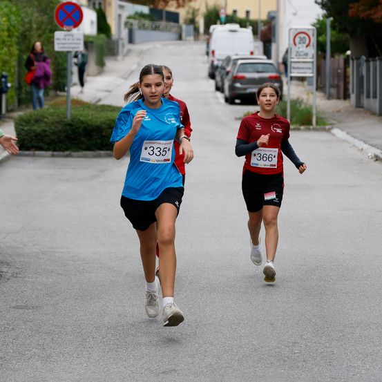 Drei Läufer nehmen an einem Straßenrennen teil. Die Läuferin vorne trägt ein blaues Shirt mit der Nummer 335, die hinter ihr laufende Läuferin trägt ein rotes Shirt mit der Nummer 366. Im Hintergrund befinden sich geparkte Autos und ein Schild.