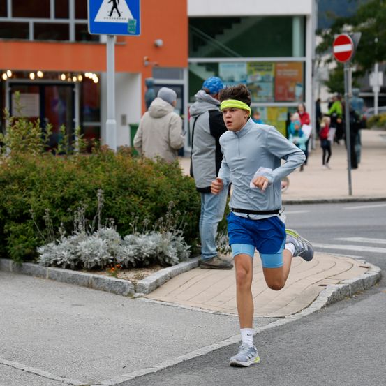 Ein junger Mann läuft auf der Straße in einer blauen Kapuzenjacke und Shorts und hält einen Plastikbeutel. Dahinter stehen zwei Personen und beobachten.