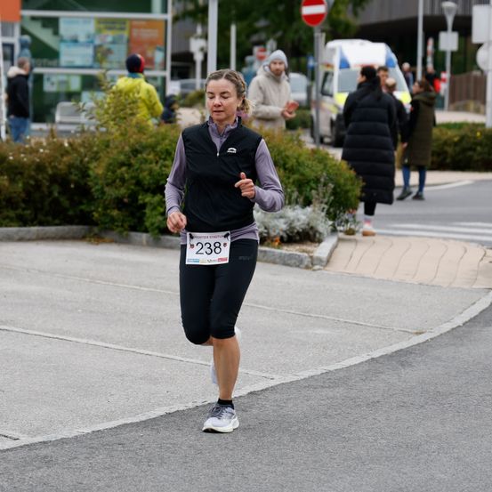 Eine Frau mit einer Weste mit der Nummer 238 läuft auf der Straße einen Marathon. Hinter ihr stehen mehrere Menschen und schauen zu.