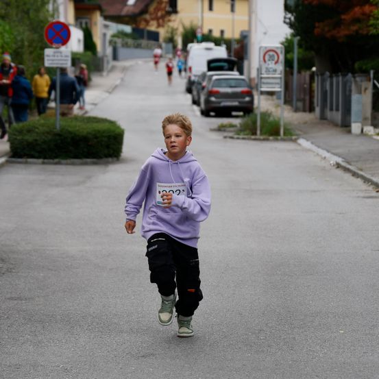 Ein junger Junge läuft auf einer Straße mit der Startnummer 1002, während andere im Hintergrund an einem Rennen teilnehmen.
