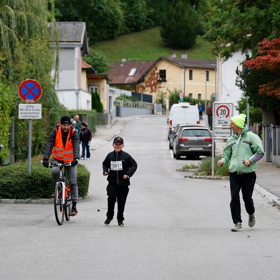 Menschen laufen auf einer Straße mit einer Geschwindigkeitsbegrenzung von 30 km/h. Ein Radfahrer ist links, und ein Läufer ist rechts. Bäume und Häuser befinden sich an den Seiten.