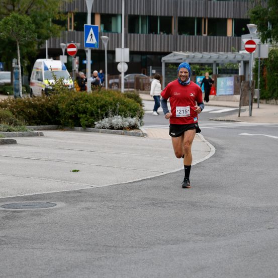 Ein Mann läuft auf einer Stadtstraße beim Marathon. Er trägt ein rotes Shirt mit der Nummer 215. Dahinter sind ein Krankenwagen und Fußgänger zu sehen.
