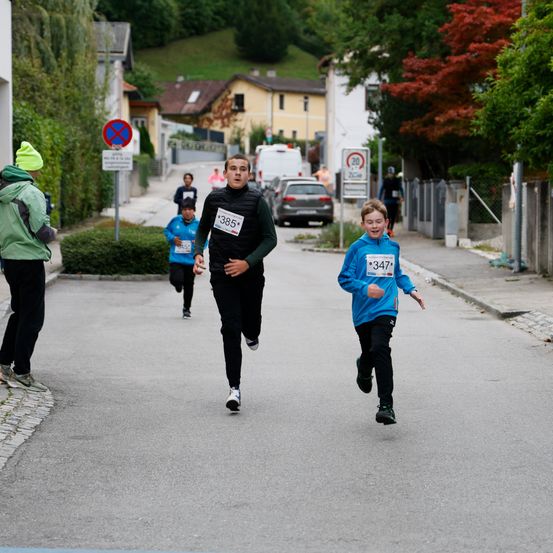 Ein Junge in einem blauen Shirt mit der Nummer 347 läuft auf der Straße. Dahinter läuft ein Junge in einem schwarzen Shirt mit der Nummer 385. Dahinter beobachtet ihn ein Mann in einem grünen Hut.