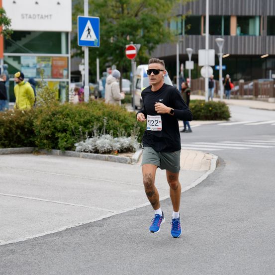 Ein Mann mit einer schwarzen Nummer 22 auf dem Shirt läuft einen Marathon auf einer Straße mit Büschen und einem Zebrastreifenschild.