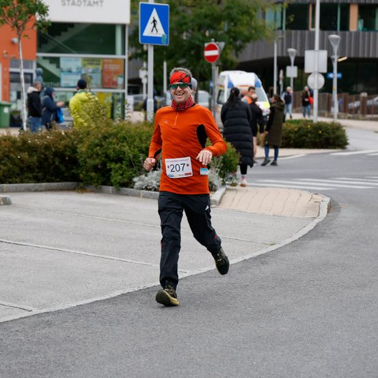Ein Mann mit Sonnenbrille und rotem Stirnband läuft einen Marathon. Er trägt die Startnummer 207. Hinter ihm sind mehrere Menschen, die auf dem Bürgersteig gehen.