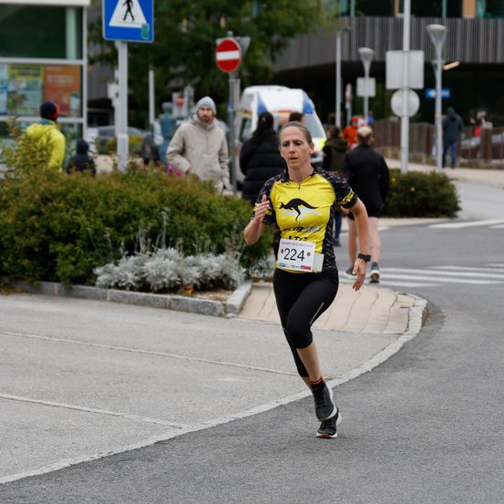 Eine Frau in einem gelben und schwarzen Shirt läuft auf der Straße, ihre Nummer 224 sichtbar. Menschen und Grünpflanzen sind im Hintergrund.