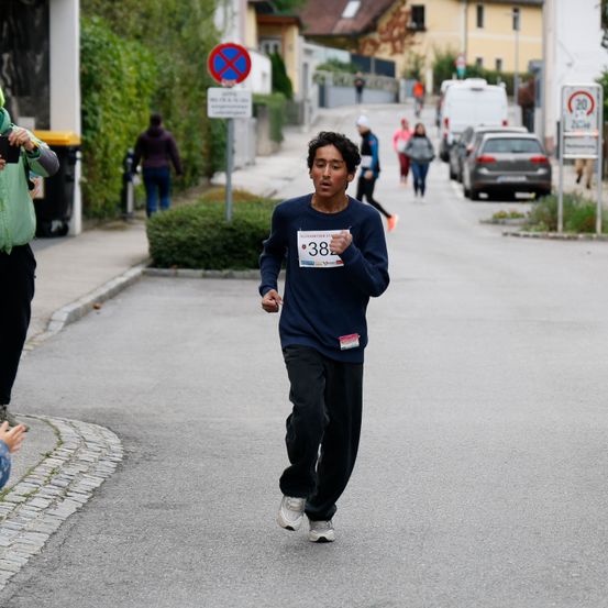 Ein junger Läufer mit der Nummer 38 auf seinem Shirt läuft eine Straße hinunter, hinter ihm laufen ein paar Leute. An der Straße sind Autos geparkt.