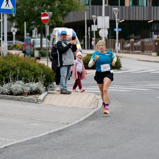 Eine Frau läuft einen Marathon in einem blauen Shirt und schwarzen Shorts. Zuschauer beobachten sie vom Bürgersteig aus.