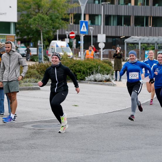Teilnehmer laufen in einem Marathon, tragen Turnschuhe und Jacken. Eine Frau vorne hat die Nummer '233' auf ihrem Shirt. Zuschauer beobachten von der Seite. Dahinter stehen Bäume, Büsche und Gebäude.