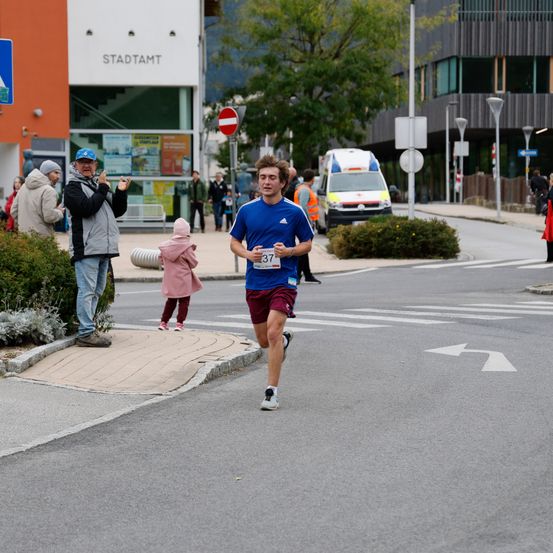 Läufer Nummer 37 joggt auf der Straße, während Leute auf dem Bürgersteig stehen, darunter ein Mann in einer grauen Jacke. Hinter ihm steht ein Schild mit der Aufschrift Stadtamt und ein geparkter Krankenwagen.