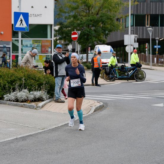 Eine Läuferin mit der Nummer 206 auf ihrem Trikot joggt auf einer Straße, wobei Menschen am Bürgersteig stehen. Ein blaues Fahrrad ist hinter einem Mann in einer orangefarbenen Weste geparkt.