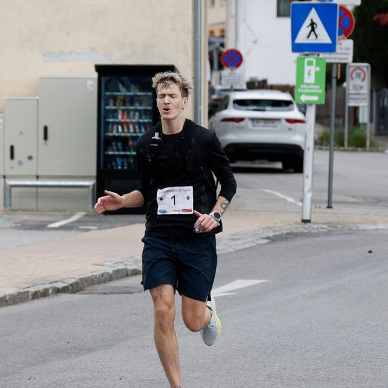 Ein Mann in einem schwarzen Shirt mit der Nummer 1 läuft auf einer Straße, ein weißer Wagen ist geparkt, und ein Fußgängerüberweg-Schild ist sichtbar.