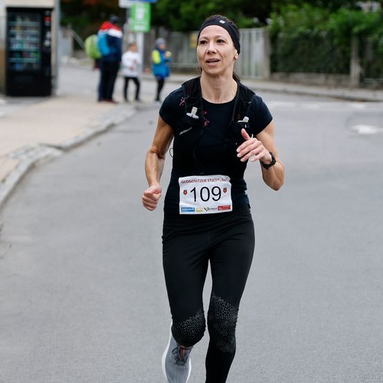 Eine Frau mit der Nummer 109 auf ihrem Shirt läuft auf einer Straße. Leute stehen im Hintergrund. Ein Verkaufsautomat ist links sichtbar.