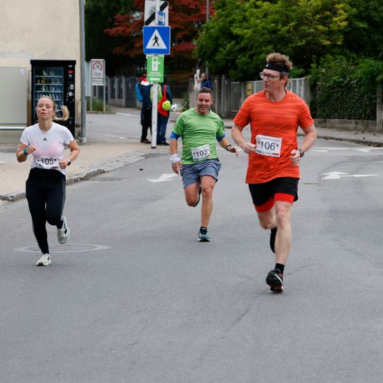 Drei Läufer nehmen an einem Marathon teil. Die Frau trägt ein weißes Shirt, der Mann in Grün trägt die Nummer 103 und der Mann in Orange trägt die Nummer 106.