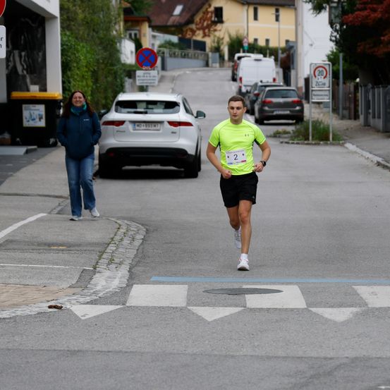 Ein Mann in einem gelben Shirt mit der Nummer 2 läuft auf der Straße. Eine Frau in einer blauen Jacke steht auf dem Bürgersteig und lächelt. Dahinter stehen geparkte Autos und ein Gebäude mit einem Mülleimer.