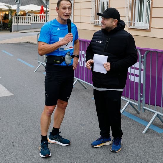 Zwei Männer stehen im Freien; einer hält ein Mikrofon, der andere ein Papier. Hinter ihnen steht eine Metallbarriere mit einem lila Banner. Hinter der Barriere sind Gebäude und eine Terrasse mit Tischen und Sonnenschirmen zu sehen.