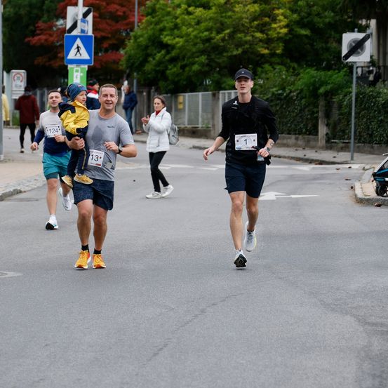 Eine Gruppe von Läufern nimmt an einem Marathon teil. Ein Mann hält ein Kind auf der Schulter, während er läuft. Ein anderer Läufer ist in Führung.