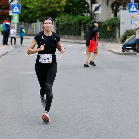 Eine Frau mit der Startnummer 109 läuft auf der Straße bei einem Marathon, trägt ein schwarzes Outfit und Turnschuhe. Hinter ihr trägt ein Mann eine rote Handtasche. Bäume und Büsche sind im Hintergrund.