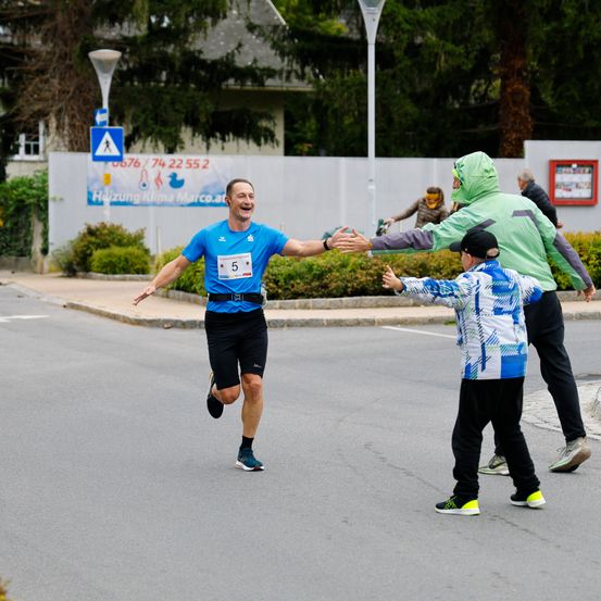 Ein Mann läuft einen Marathon. Er wird von einem Mann in einer grünen Jacke gratuliert.