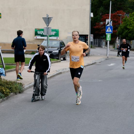 Ein Mann mit Brille läuft auf der Straße, während ein Junge einen Roller fährt. Zwei andere Leute rennen hinterher. Ein Gebäude mit Schildern und ein Auto befinden sich am Straßenrand.