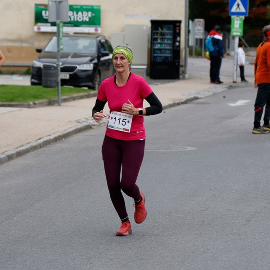 Eine Frau in einem pinkfarbenen Shirt mit der Nummer 115 läuft auf der Straße, mit einem schwarzen Auto, das hinter ihr geparkt ist.