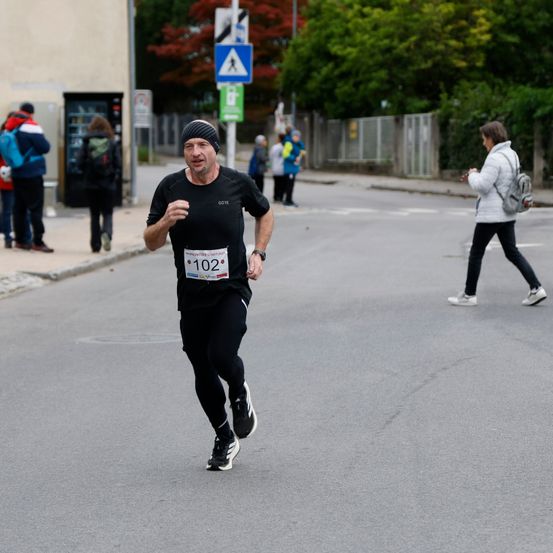 Ein Mann in einem schwarzen Shirt und schwarzen Hosen läuft bei einem Marathon mit der Nummer 102 auf seinem Shirt. Eine Frau mit einer Tasche geht im Hintergrund.