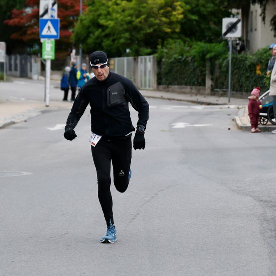 Ein Mann läuft auf der Straße mit einer Startnummer und trägt ein schwarzes Outfit, eine Brille und Handschuhe. Hinter ihm laufen Leute, und ein Kinderwagen steht in der Nähe. Ein Zaun trennt die Straße vom Bürgersteig.