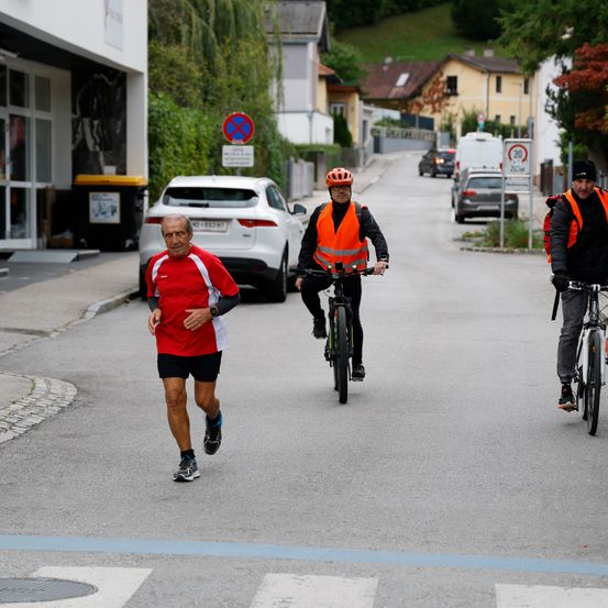 Ein Mann rennt auf der Straße, während zwei Männer auf Fahrrädern folgen. Ein weißer Wagen ist am Straßenrand geparkt.