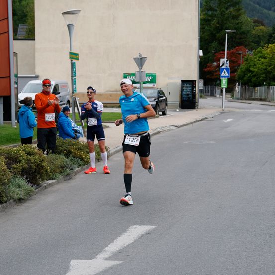 Ein Läufer mit der Nummer 125 läuft auf der Straße. Hinter ihm stehen mehrere Leute auf der Seite, von denen einer ein orangefarbenes Shirt trägt.