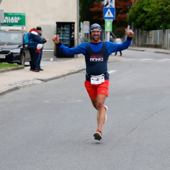 Ein Mann in einem blauen Shirt und roten Shorts läuft bei einem Marathon. Er hält die Arme in der Luft und trägt einen blauen Helm. Hinter ihm stehen zwei Personen auf dem Bürgersteig in der Nähe eines Verkaufsautomaten. Ein schwarzer Wagen ist am Straßenrand geparkt.