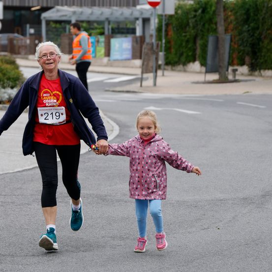 Eine ältere Frau läuft einen Marathon mit einem kleinen Mädchen, das sie an der Hand hält. Die Frau trägt ein rotes T-Shirt mit der Nummer 219. Sie sind auf einer Straße, mit einem Gebäude und einem Verkehrsschild im Hintergrund.
