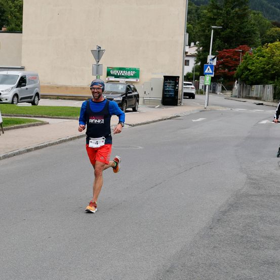 Ein Mann läuft auf der Straße mit einer Kopfband und einem nummerierten Shirt. Dahinter ist ein Van geparkt und ein Auto fährt.
