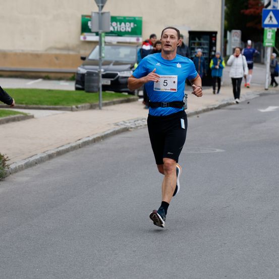 Ein Mann läuft auf der Straße mit der Nummer 5 auf seinem T-Shirt, trägt Ohrhörer und eine Wasserflasche. Hinter ihm laufen Menschen, und Autos sind geparkt.