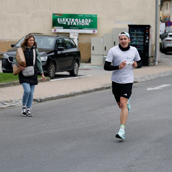 Ein Mann läuft auf der Straße in einem weißen T-Shirt und schwarzen Shorts. Eine Frau geht auf dem Bürgersteig. Dahinter steht ein schwarzer Wagen.