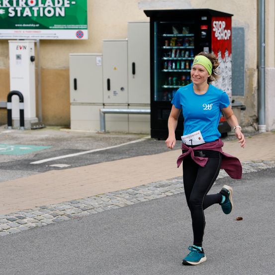 Eine Frau läuft auf einer Straße, trägt ein blaues Shirt mit der Nummer 7 und schwarze Leggings. Sie hat eine grüne Stirnband und lächelt. Hinter ihr befindet sich ein Verkaufsautomat und ein grünes Schild.