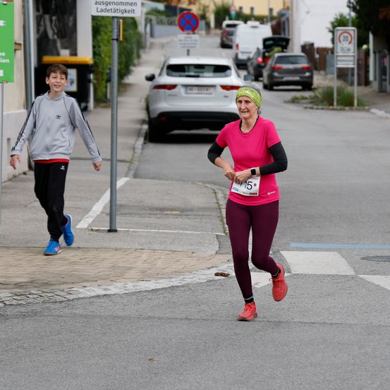 Eine Frau joggt auf einer Straße, und ein Junge läuft neben ihr. Dahinter sind geparkte Autos. Die Straße hat einen Zebrastreifen mit weißen Linien. Straßenschilder und ein Mülleimer sind auf dem Bürgersteig.