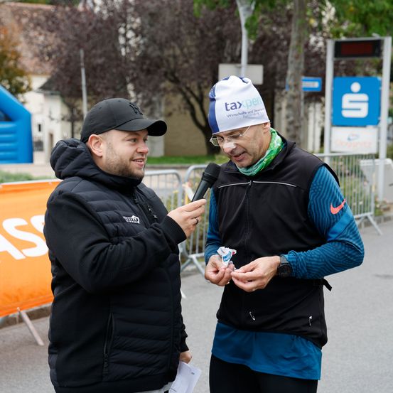Ein Mann in einer schwarzen Jacke wird von einem anderen Mann interviewt, der eine Mütze mit einem taxpen-Logo trägt. Der interviewte Mann hält einen kleinen Gegenstand in der Hand. Die Szene spielt sich im Freien in der Nähe eines Zauns ab.