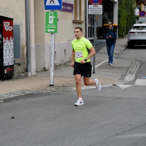 Ein Mann läuft die Straße entlang und trägt ein neongelbes Shirt, schwarze Shorts und weiße Sneaker. Eine Frau in einer blauen Jacke und Jeans geht auf dem Bürgersteig in der Nähe. Straßenschilder und ein Verkaufsautomat sind sichtbar.