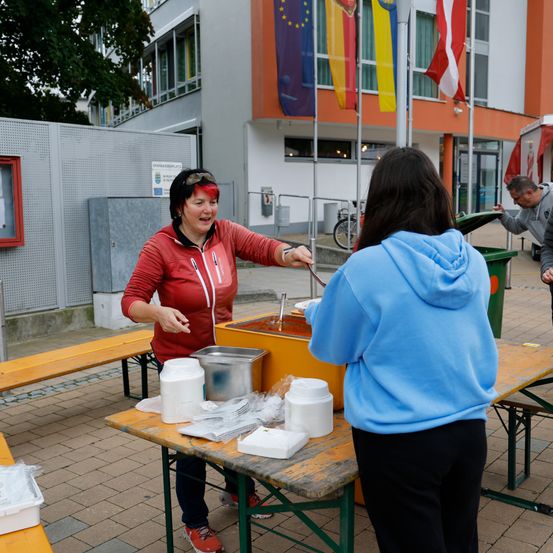 Eine Frau serviert Essen aus einem gelben Behälter an eine andere Frau vor einem Tisch vor einem Gebäude mit Flaggen.