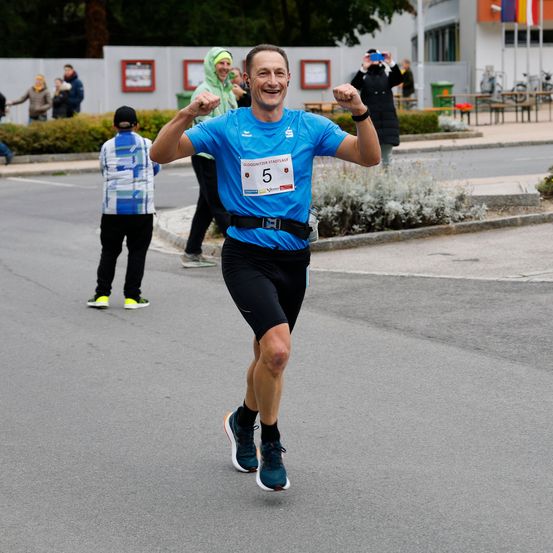 Ein Mann läuft einen Marathon mit der Nummer 5 auf seinem Shirt und feiert seinen Erfolg. Hinter ihm stehen Menschen, die zusehen und jubeln.