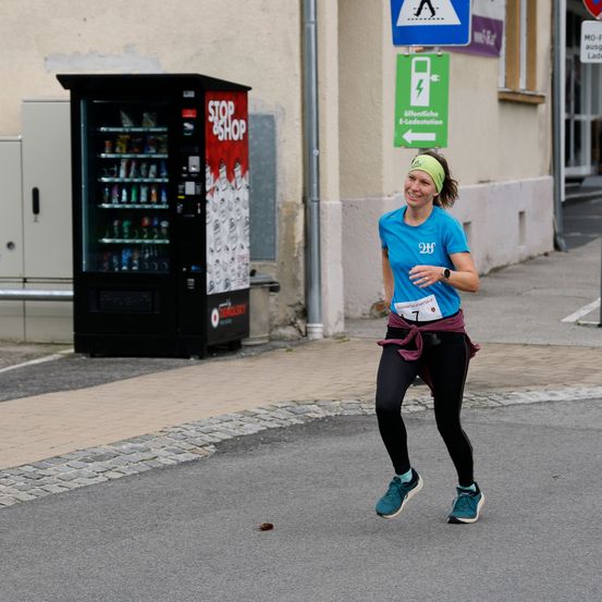 Eine Frau in einem blauen Shirt und schwarzen Leggings läuft einen Marathon auf einer gepflasterten Straße mit einem Verkaufsautomaten an der Seite.