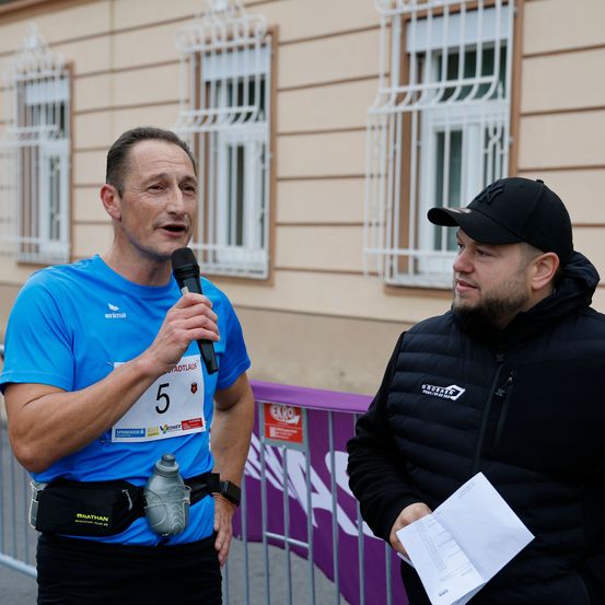 Ein Mann in einem blauen Shirt und Laufausrüstung spricht in ein Mikrofon, während ein anderer Mann in einer schwarzen Jacke zuhört und ein Papier in der Hand hält. Sie stehen vor einer Metallbarriere mit einem Gebäude mit weißen Fenstern im Hintergrund.