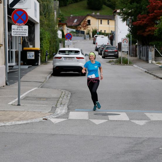Eine Frau läuft auf einer Straße und überquert einen Zebrastreifen. Auf beiden Seiten der Straße sind mehrere Autos geparkt. Straßenschilder und ein Gebäude sind auf der linken Seite sichtbar.