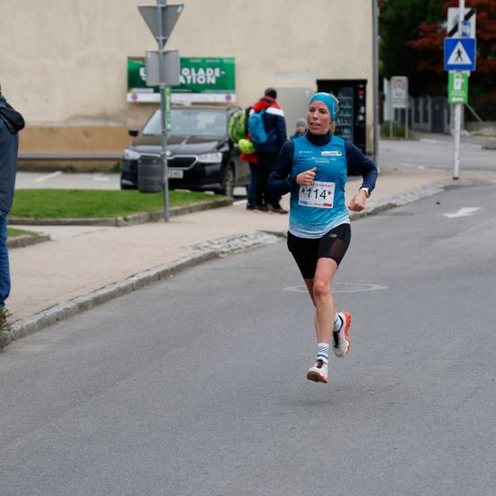Eine Frau in einem blauen Shirt und schwarzen Shorts läuft einen Marathon. Die Nummer 114 ist auf ihrem Shirt zu sehen. Hinter ihr stehen ein schwarzer Wagen und ein Mülleimer am Bürgersteig.