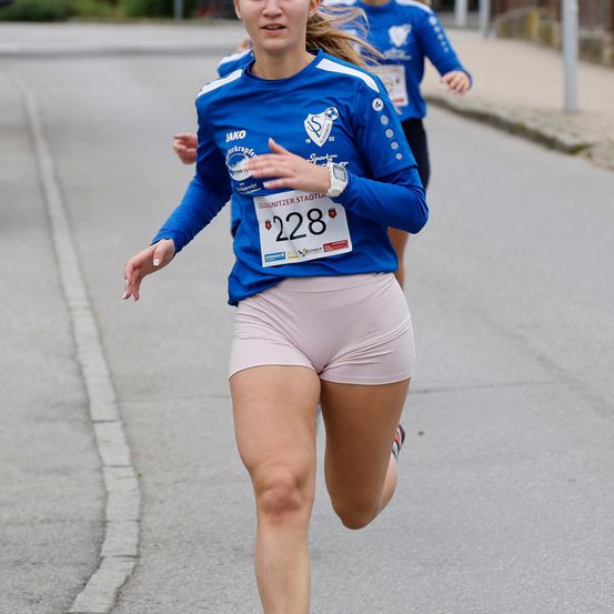 Eine Frau mit einem blauen Shirt, auf dem die Nummer 228 steht, läuft auf der Straße. Hinter ihr läuft eine weitere Person. Der Hintergrund ist verschwommen.
