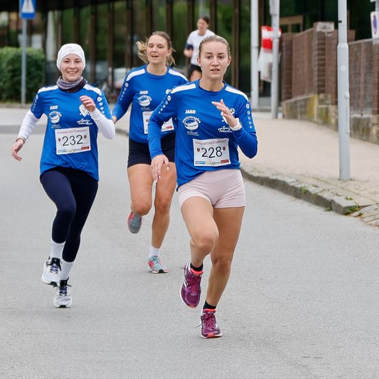 Drei Läuferinnen nehmen an einem Marathon teil, tragen einheitliche blaue Trikots mit den Nummern 232, 228 und 248. Sie laufen die Straße hinunter, wobei die mittlere Läuferin führt. Dahinter läuft ein Mann in die entgegengesetzte Richtung.