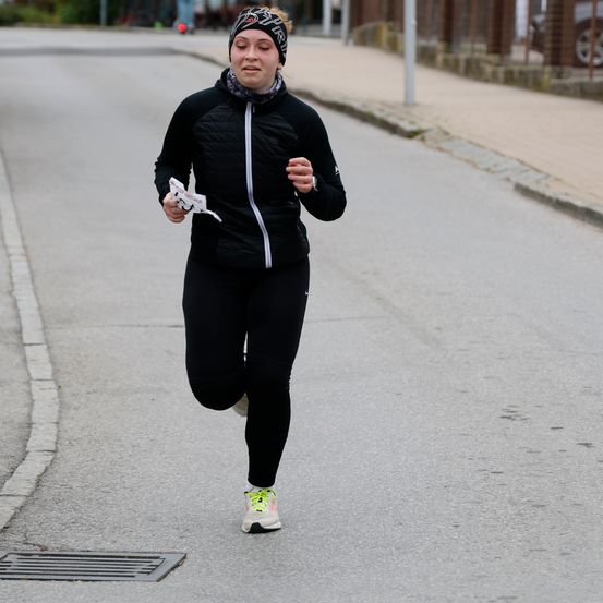 Eine Frau läuft auf einer Straße und hält ein kleines Stück Papier in der Hand. Sie trägt eine schwarze Jacke, Hose und Turnschuhe.